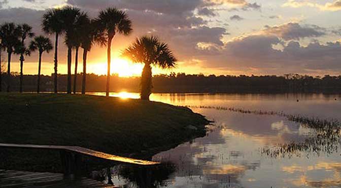 Sunrise over a tranquil lake with silhouettes of palm trees and a reflection of the sun on the water near Rollins College.