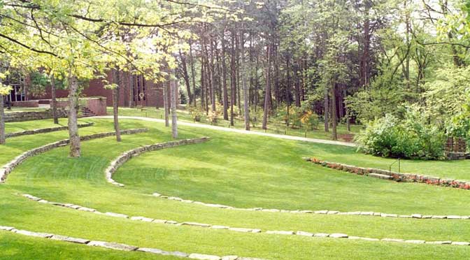 Lush green terraced garden with curved stone-edged grass paths leading through a wooded Doane College park area.