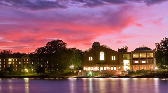 Twilight view of a modern Saint Norbert College building with large windows and a curved roof beside a lake, under a pink and blue sky.