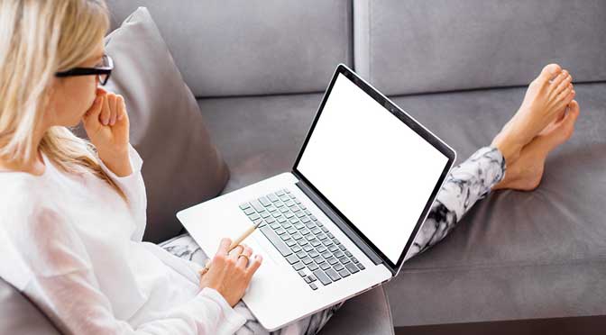 Woman in glasses and casual attire using a laptop with a blank screen to research ways to pay less for college, sitting on a gray sofa with her feet up.