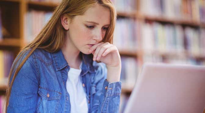 A focused young woman in a denim jacket using a laptop at a library table, researching how to decide between two universities, surrounded by books.