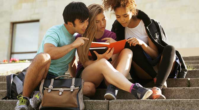 Three students from one of the top 50 colleges in US News Best College Rankings, sitting on steps outdoors, studying together from a textbook.