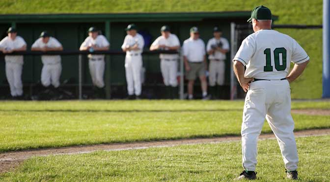 A baseball coach in a white and green uniform stands on the field with hands on hips, embodying what college baseball coaches want in terms of discipline and focus, while players and staff lean against the dugout fence in the background.